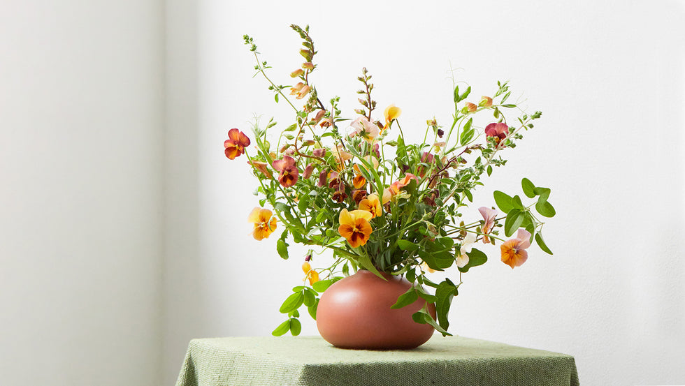A flower arrangement with small orange, yellow, and red blooms in a round, reddish-brown vase placed on a green cloth-covered table against a plain white background.